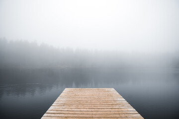 Fototapeta premium Wooden Pier Extending into Thick Morning Mist over a Calm Lake, Moody November Landscape in Soft Natural Light
