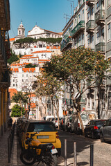Picturesque Lisbon street scene featuring vibrant cars, traditional buildings, leafy trees, and a scenic view of a historic hilltop church under a clear blue sky. Lisbon, Portugal - 11 June 2025