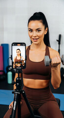 Fototapeta premium Fit woman filming workout video with dumbbell and smartphone on tripod