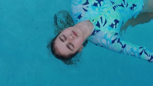 Happy Child relaxing in blue swimming pool. Top View Girl floats on her back in clear, bright blue water. His eyes are closed in peaceful relaxation. Serene, calm mood. Tranquility and summer.