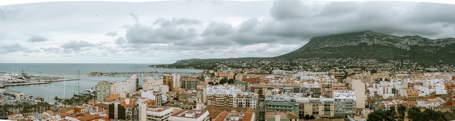 A panoramic view of Denia, Spain, showcasing the coastal cityscape and mountainous backdrop under cloudy skies.