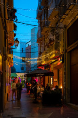 Naklejka premium Cozy European street scene at dusk with string lights, people dining outdoors, and vibrant ambiance. Perfect for travel, nightlife, and urban lifestyle inspiration.