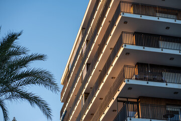Obraz premium Low angle view of modern apartment building balconies in Valencia, Spain, with warm sunset light and palm tree against clear blue sky for urban lifestyle concepts.