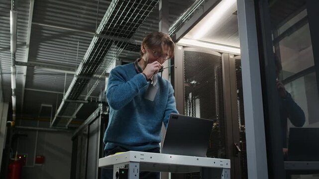 Medium shot of professional Caucasian male technician coordinating through walkie talkie while operating laptop in data center aisle with server racks