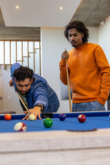 Indian friends playing pool in home rec room, leaning and lining up shot on blue-felt table
