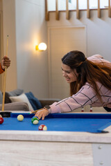 Fototapeta premium Asian adult woman and friend leaning over blue pool table with balls at home, wearing smartwatch