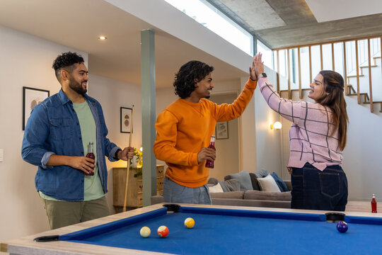 Diverse friends high-fiving while holding bottled drinks behind blue pool table in basement
