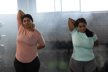 African American female friends stretching in gym beside dumbbell rack, wearing pink and blue tops