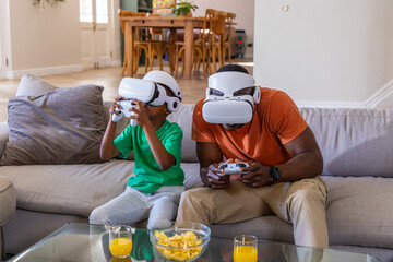 African American father and son sitting in living room wearing VR headsets, holding controllers © wavebreak3
