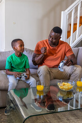 African American father-son playing games on gray sofa, holding white controllers, juice and chips © wavebreak3