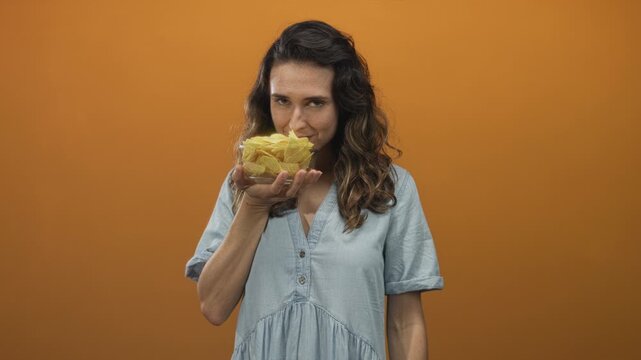 Woman holds bowl of potato chips close to her nose in orange studio; craving indulgence appetite satisfaction.