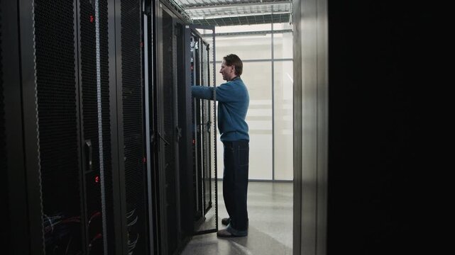 Side view full shot of Caucasian male network engineer approaching server rack, connecting network cable and installing server hardware in modern data center corridor