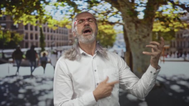 Man with long grey hair and red glasses sings with hand to chest and other hand gesturing on a street by trees and buildings; passion expression.