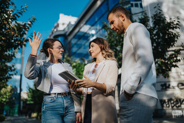 Three professionals in business casual discuss ideas outdoors. They stand on a sunny city street,...