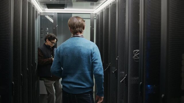Medium tracking shot of network engineer closing server rack after maintenance, shaking hands with Asian colleague working on laptop before leaving data center corridor