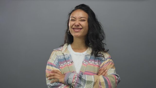 Young hispanic woman with arms crossed and squinting smile wearing patterned jacket in gray studio; playful confidence.