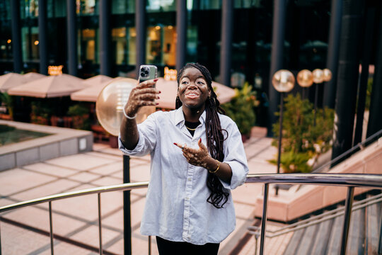 Excited young woman with vitiligo pointing at phone screen during video chat, laughing with joy and expressing emotional connection through digital self-representation