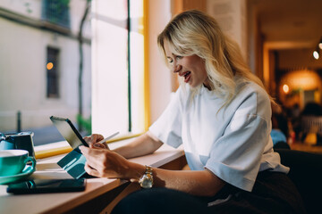 Laughing woman using stylus on tablet while working remotely at window seat, symbolizing creative...