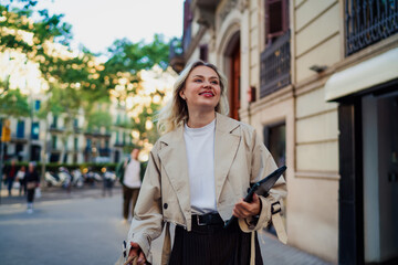 Obraz premium Blonde woman confidently walking down city street holding tablet and handbag. Image illustrates mobile productivity, personal freedom, and tech-driven professional lifestyle