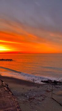 Cinematic 4K vertical sunset over the Mediterranean coast of Varazze on the Ligurian Sea in Italy, with dramatic orange sky reflecting on calm waves and rocky shoreline.