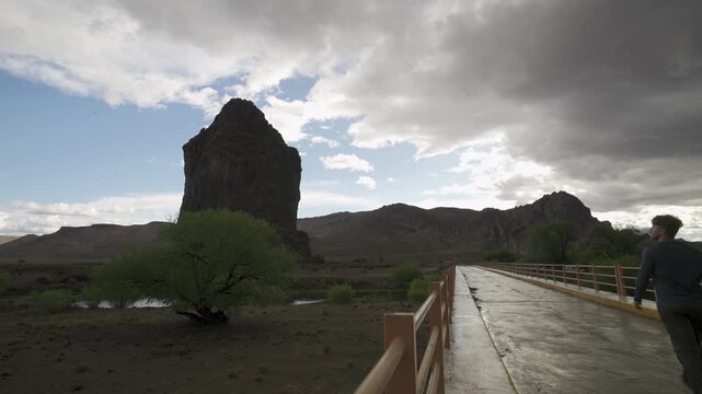 Man crossing bridge toward Piedra Parada rock formation in vast Patagonia steppe on a cloudy afternoon. Chubut, Patagonia, Argentina