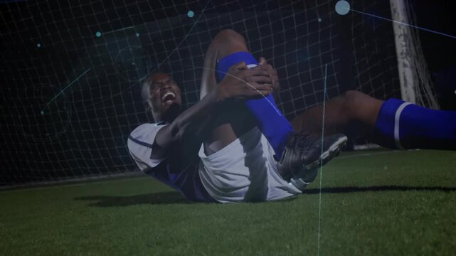 Male soccer player lying by goal net clutching knee from pain, camera pulling back revealing smile