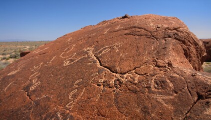 Displaying large sandstone boulder bearing carved petroglyphs on arid plain with winding dirt track