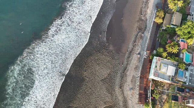 Drone flying along volcanic black sand beach with palm trees, ocean waves and coastal buildings in El Tunco, La Libertad, El Salvador, Central America.