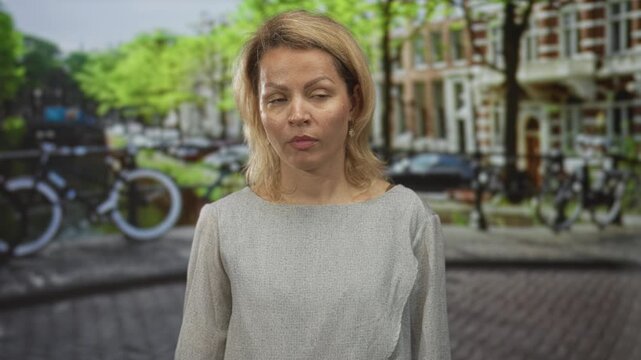 Woman stands with bare face on a busy city street lined with brick buildings and parked bicycles; apathy.
