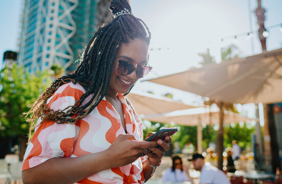 Smiling woman in patterned shirt using smartphone with both hands outdoors, representing digital engagement, social connection, and seamless integration of mobile technology.