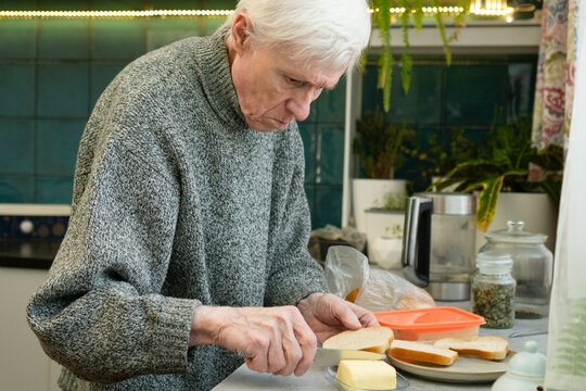 A senior citizen spreads butter on bread to make sandwiches. The image shows self-sufficiency and simple meal preparation in old age. Elderly man handling food independently.