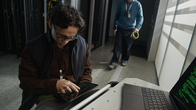 Tracking shot of two network engineers performing server configuration and cable installation in data center, technician coding on tablet and laptop while colleague connecting cable to rack equipment