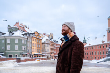 A man in winter attire stands smiling in Warsaw’s Castle Square, surrounded by the snow-dusted, colorful facades of the historic Old Town.