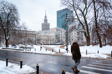 A man crossing a snow-lined street in Warsaw, where the bare branches of winter trees frame a view of the iconic Palace of Culture and Science rising behind a modern glass skyscraper.