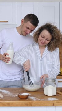 Loving couple preparing breakfast together in their pajamas on a cozy morning. The woman whisks batter in a glass bowl while her smiling husband hugs her, holding a bottle of milk