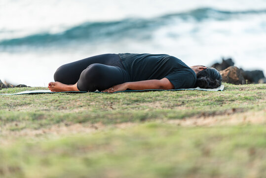 Woman practicing a reclining supine twist yoga pose on a mat by the ocean
