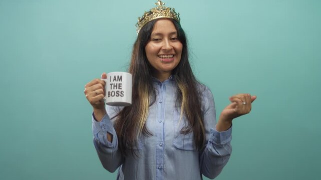 Woman wearing a crown and light blue shirt holding a mug that reads i am the boss while pointing at the camera in a teal studio; confidence authority.