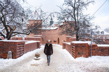 A man walks along a snow-covered path toward the Warsaw Barbican, a historic red-brick semi-circular fortified gate that serves as a gateway between the Old and New Town.
