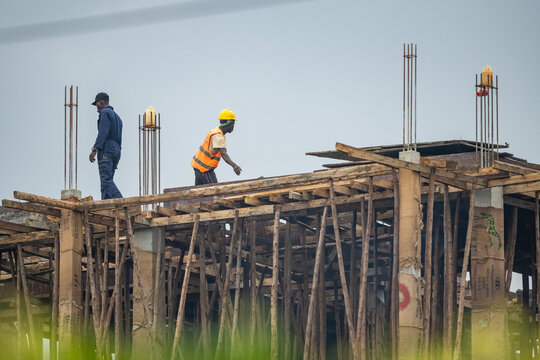 Construction workers wearing safety helmets working on a building site near Entebbe, Uganda. African builders installing wooden formwork on a structure under clear blue sky.