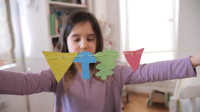 Little girl smiling at the camera while proudly holding up a string of colorful handmade paper bunting flags, showcasing her creativity and diy home decoration efforts