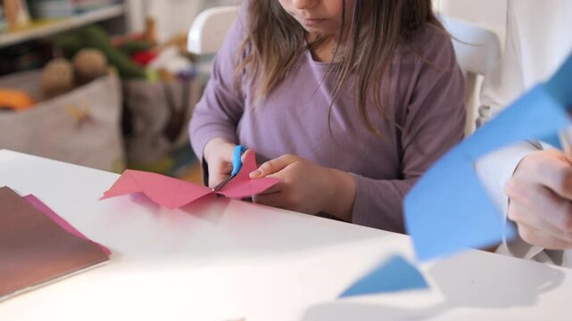 Young child focusing on cutting folded pink paper with blue handle scissors at a white table, engaging in a school project or home craft activity with concentration and developing fine motor skills