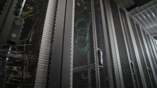 Panning view of server rack rows with connected ethernet cables and blinking indicators in modern data center corridor, low angle shot showing internet infrastructure and data storage hardware