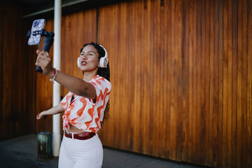 Confident woman vlogging with phone on stabilizer while talking and wearing headphones, content...