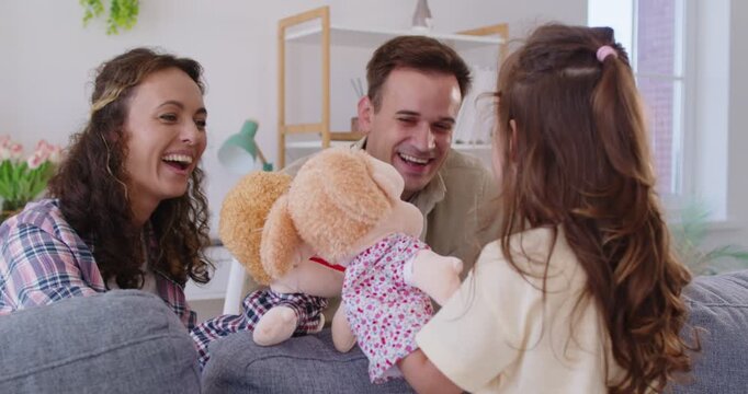 Happy playful family at home play puppet theater show with child girl. Parents and daughter animate plush characters on sofa, laughing together indoors. Family bonding and leisure concept.