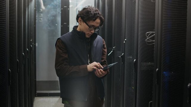 Medium portrait shot of Asian network engineer standing between server racks in data center corridor holding digital tablet and smiling confidently at camera