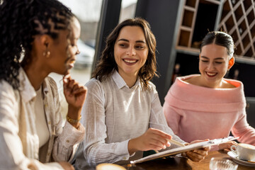 Fototapeta premium Three cheerful diverse business women laughing while reviewing project documents in a cafe.