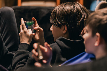 Two adolescent boys engaged with smartphones while seated on a couch, one holding a phone and the other with a hand raised, indoor setting with soft lighting © Emvats
