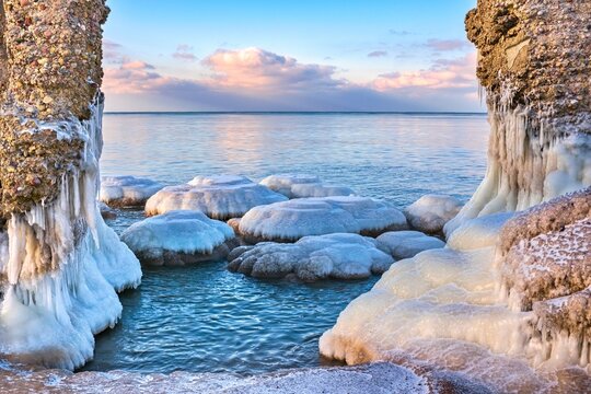 Unique ice formations and icicles on coastal ruins at winter sunset. Beautiful frozen sea landscape with round ice blocks and calm water under a pink sky. Arctic nature background.