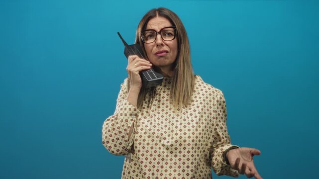 Woman holds handset to ear in studio with blue backdrop, wearing glasses and patterned blouse; frustration impatience.