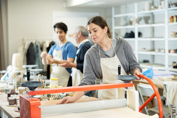 Young woman rolls raw clay with mechanical slab roller machine in pottery workshop